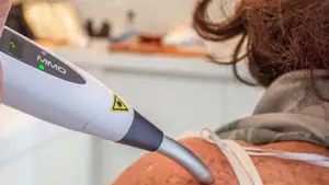 isioterapeuta da Fisiome realiza atendimento domiciliar em Boa Viagem, Recife, utilizando um equipamento de laserterapia para fotobiomodulação no joelho de uma paciente confortavelmente acomodada em sua sala de estar.