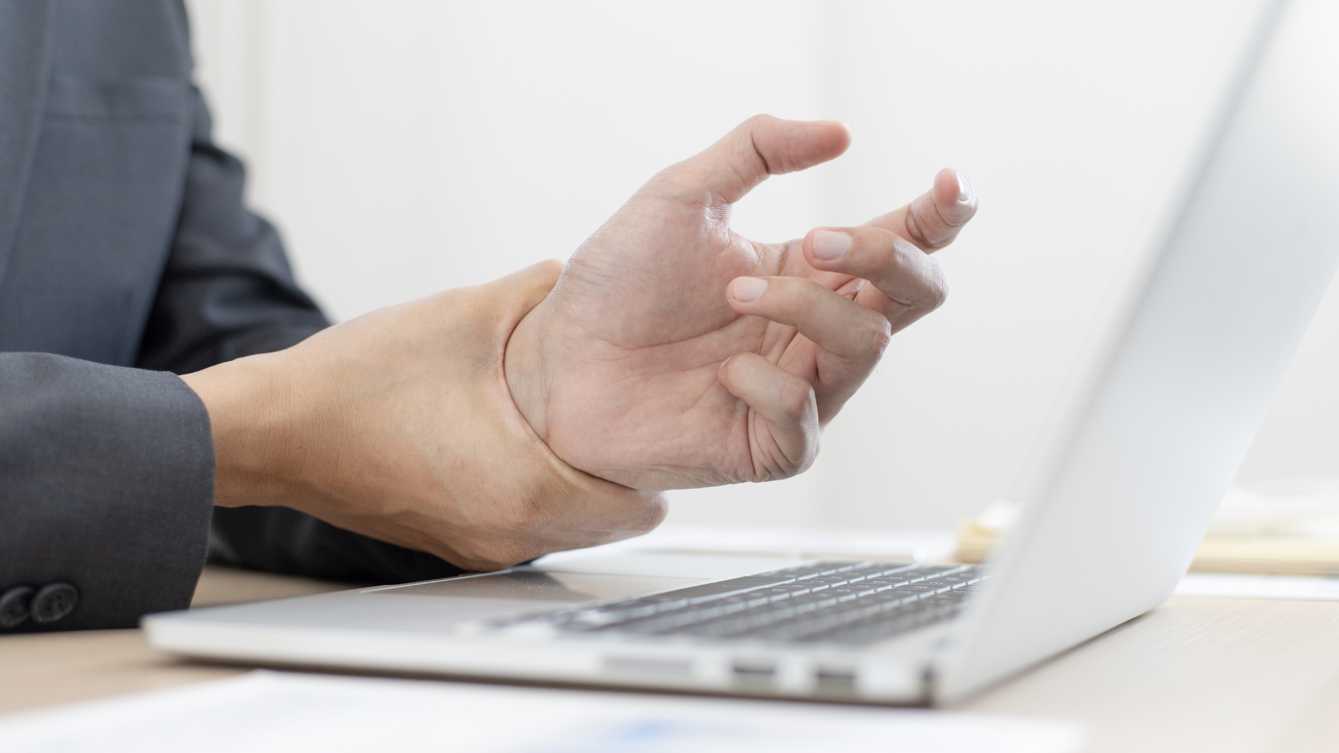 Close-up das mãos de um profissional vestindo um terno cinza, segurando e massageando um pulso dolorido com a outra mão. A mão afetada está curvada e tensa na frente de um laptop aberto, sugerindo dor por esforço repetitivo (LER/DORT) ou tendinite.